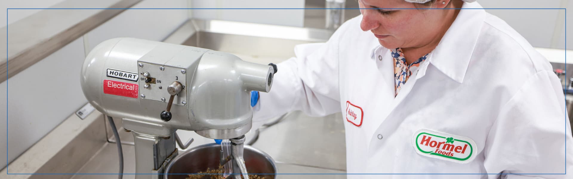 A food science worker using a commercial Hobart mixer in a clean processing lab, wearing protective gloves and a Hormel Foods lab coat.