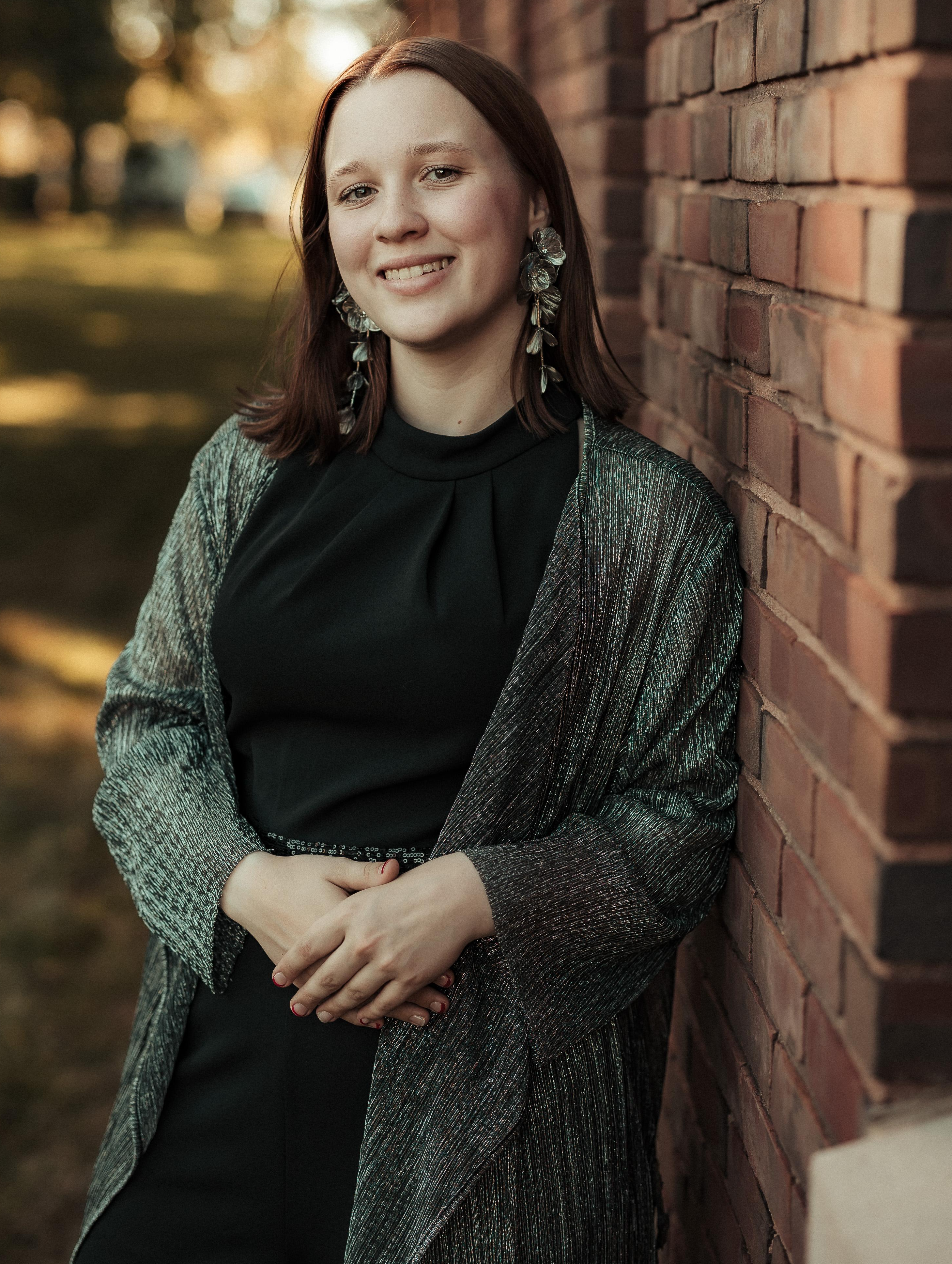 Posed photo of Katherine Eliason, outside leaning against a brick wall. She is smiling and has her hands folded in front of her.